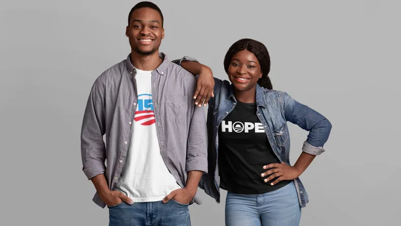 A man with dark skin tone smiles with his hands in his pockets while wearing a "15" year Obama Alumni shirt. A woman with dark skin tone smiles with her arm on the man beside her, while wearing an Obama HOPE shirt. 