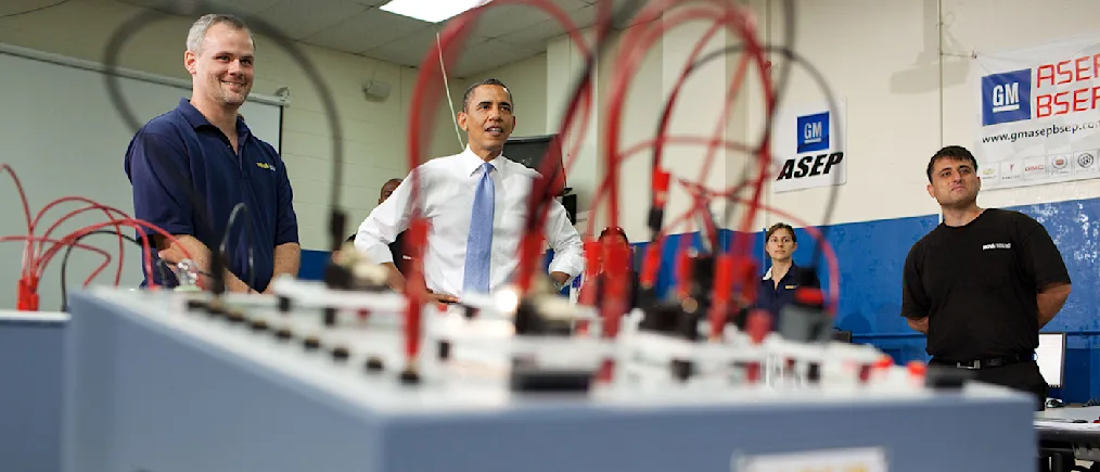 President Obama stands with a group of people with various skin tone. The camera captures a red and black wired switchboard out of focus.