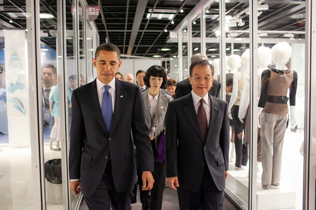 President walks down a hallway with clear cases filled with maniquens with a group of older individuals with various skin tones in buisness attire 