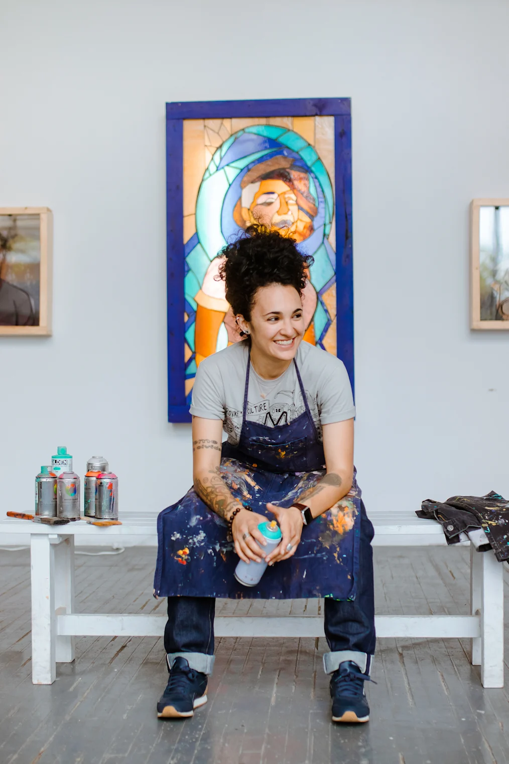 Sam Kirk is sitting on a white bench in her art studio. On the bench are several cans of spray paint. She is looking to her left and laughing. Sam has a medium light skin tone and long dark curly hair. Her hair is pulled to the top of her head. She is wearing a t-shirt, a painter's smock/apron, dark colored jeans, and sneakers. She is holding a can of spray paint. On the wall behind her is a stained glass art piece.