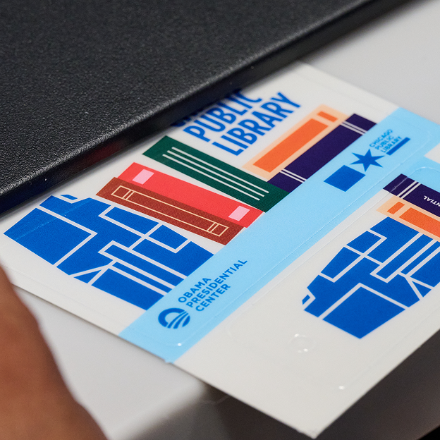 A close-up, high-angle photograph shows a white library card tucked into the pocket of a dark grey or black textured folder or book cover. The card features bold blue text at the top that reads "PUBLIC LIBRARY". Below the text, colorful abstract book icons in green, orange, red, and dark blue sit in a diagonal row. A bright blue diagonal band stretches across the card with the text "OBAMA PRESIDENTIAL CENTER" next to a white circular logo. To the right, a blue star and the text "CHICAGO PUBLIC LIBRARY" are visible above a blue pattern that looks like a map or grid. In the bottom-left corner, a person's hand with a medium-dark skin tone holds the corner of the paper.