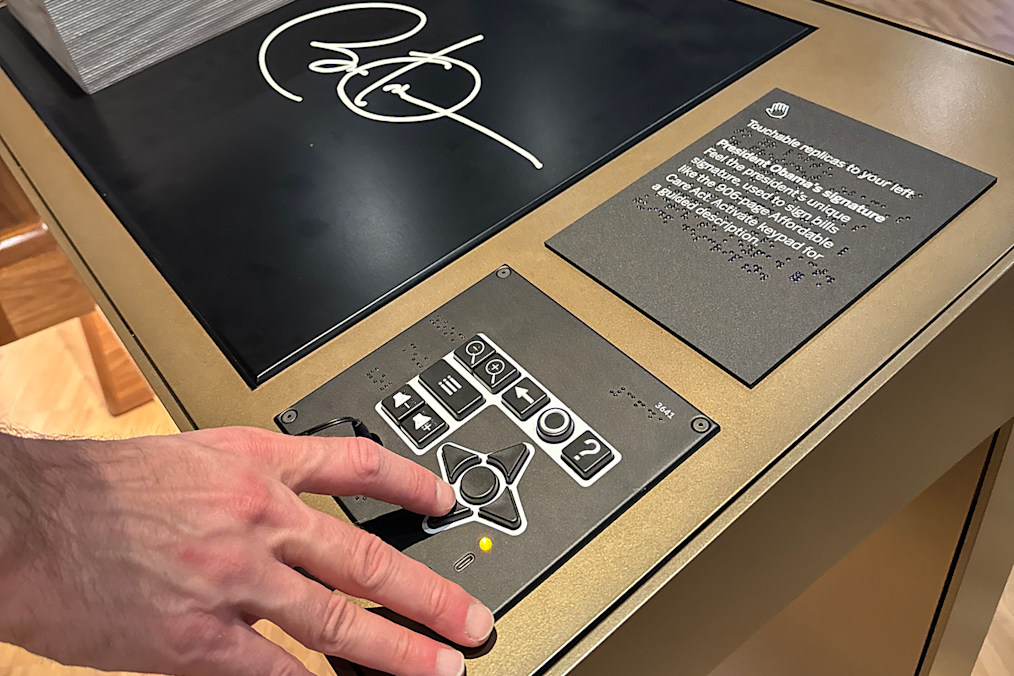 A hand with light skin tone rests on the arrow pad of a UXP-D, mounted below and to the right of a raised plaque of President Obama's signature on the sloped surface of a museum interactive.