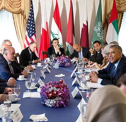 President Obama wearing a black suit with a white button-up shirt sits at a long blue table amongst a group of individuals with different skin tones in a room with white walls that have four flags in front of it. 