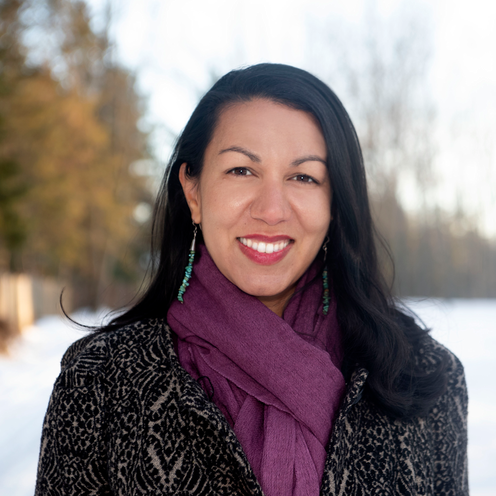 Kesha Ram Hinsdale, a woman with a light skin tone and long black hair, is smiling at the camera. She is outside and wearing a black coat with a purple scarf.