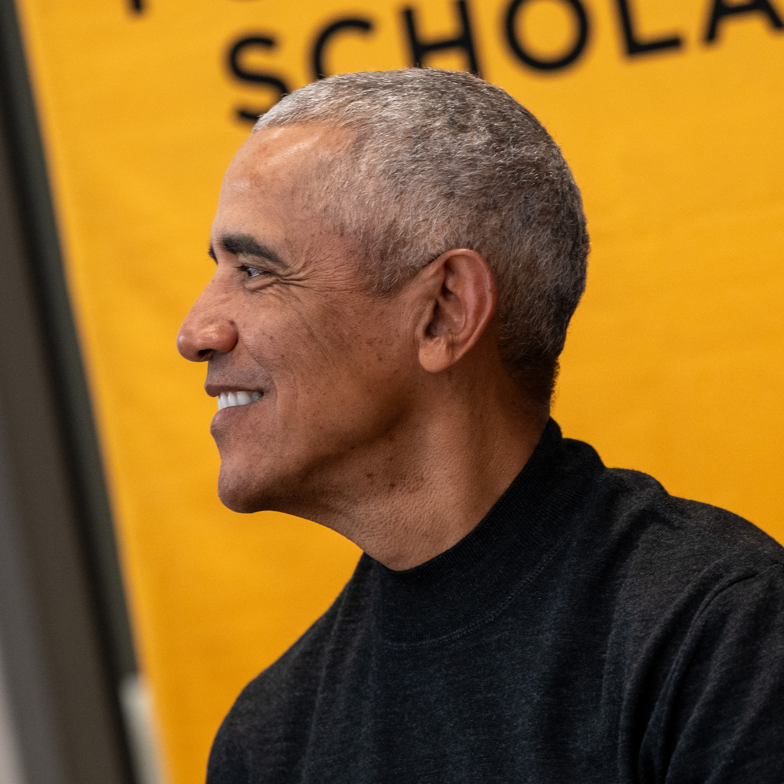 A side profile of President Obama smiling. A yellow sign in the background reads, “Scholars.”