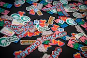 A group of colorful stickers sit on a table. Some are of The Obama Foundation logo and others say, “Obama Scholars,” “Chicago,” and “When they go low we go high.”
