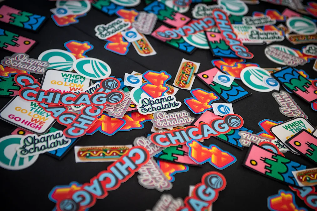 A group of colorful stickers sit on a table. Some are of The Obama Foundation logo and others say, “Obama Scholars,” “Chicago,” and “When they go low we go high.”