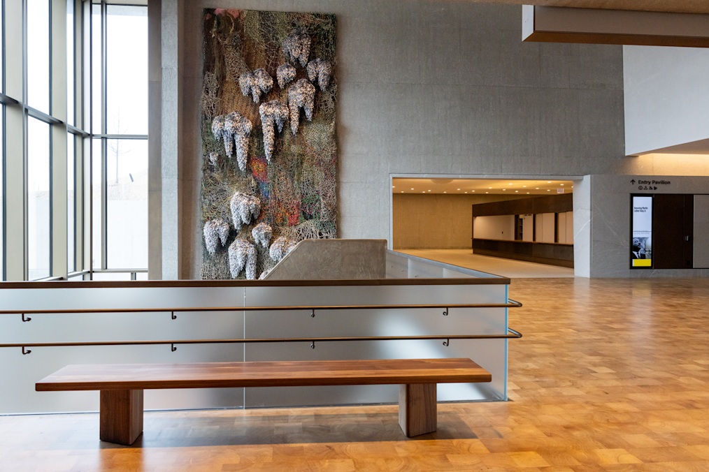 A wooden bench sits in front of a frosted glass railing overlooking a large fiber art wall hanging and a lobby area. The art in the background is This Land, Shared Sky, a new art installation. The installation is a large, vertical, mixed-media textile artwork featuring dense, net-like webbing in earthy green and brown tones. Bunches of white and gray fiber clusters extend outward from the surface, creating a textured, dimensional effect.