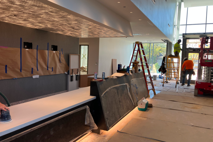 A photograph of construction on the Lobby of Home Court at the Obama Presidential Center. The picture features an image of the Welcome Desk in the Lobby and several construction workers working on the front entrance of the building.