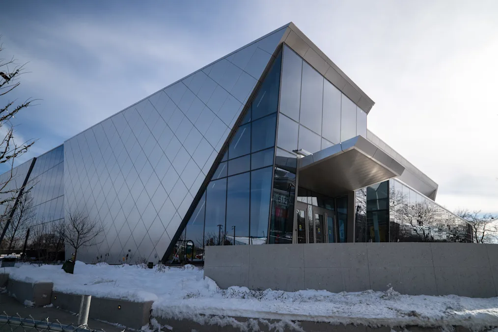 An image of the exterior of Home Court. The building is dark steel and grey, made to reflect the netting of a basketball net. Near the building, snow sits on the ground.