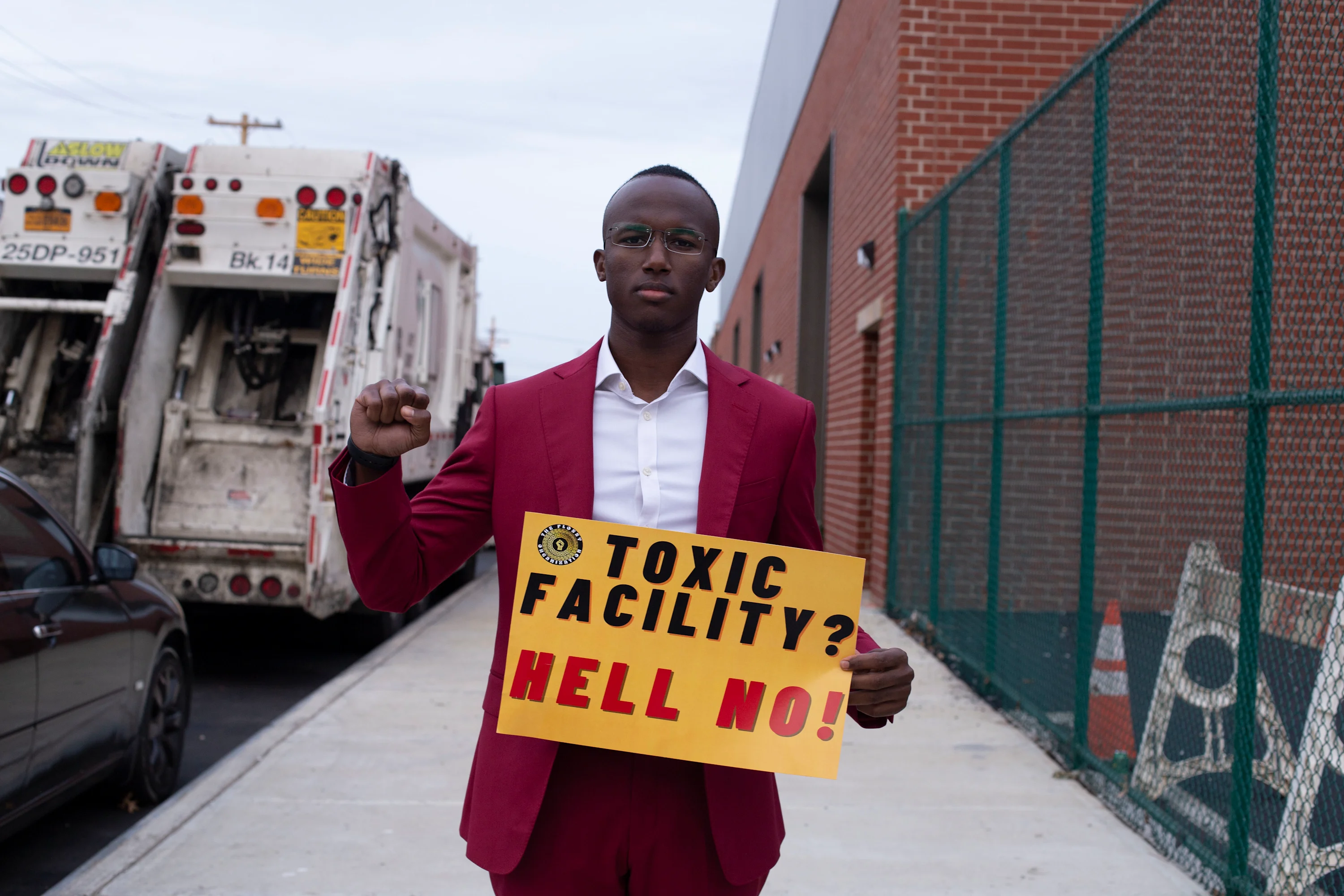 Jibreel Jalloh, a Black male with a dark skin tone, holds a sign that reads, “Toxic Facility? Hell No.” He is wearing a burgundy suit.