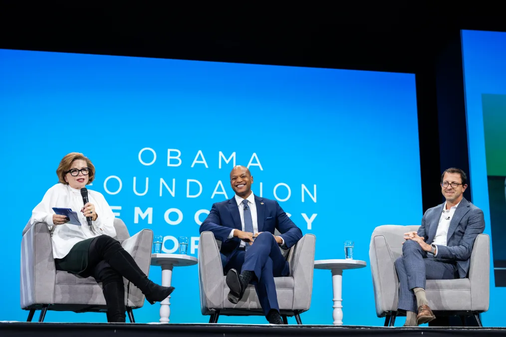 Valerie Jarrett sits on a panel alongside Maryland governor Wes Moore, a Black man with a medium skin tone and no hair, and Ian Bassin, a man with a light skin tone. The screen behind them reads, “Obama Foundation Democracy Forum.”
