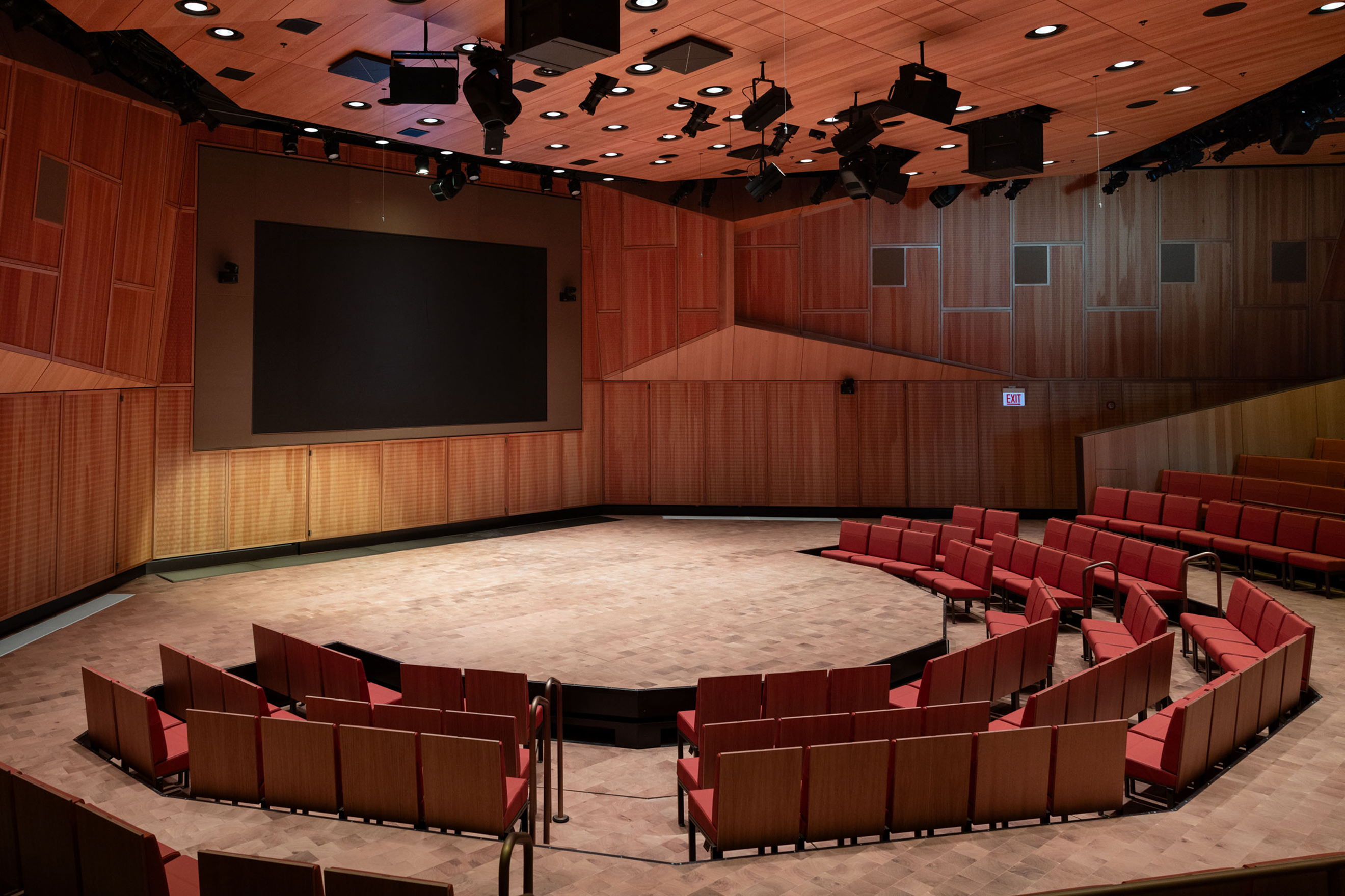 A rendering of the Elie Wiesel Auditorium located in the Forum Building of the Obama Presidential Center. The stage is a large circle surrounded by red chairs. Behind the stage is a large screen. The walls of the auditorium have wood paneling.