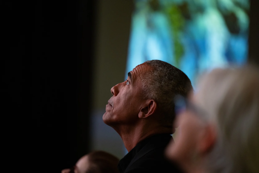 President Obama tilts his head upwards along with out-of-focus onlookers. A digital screen shows something out of focus behind him.