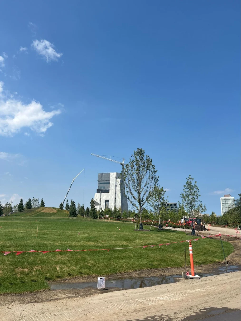 An image of the Obama Presidential Center landscape with the Obama Presidential Center Museum in the background. In the front of the image are planted trees surrounded by soil and dirt. In front of the trees and dirt are flags and construction cones. In the background is the Museum Building with a crane on top of the building. Next to the trees is a dirt path. It is daylight outside and the sky is blue.