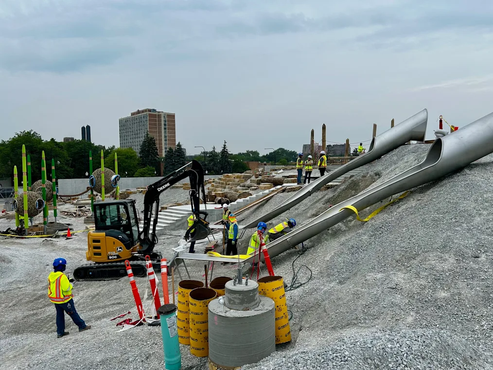 An image of construction on the Obama Presidential Center Playground. In the image, several construction workers are building two large silver slides. They are surrounded by gravel and dirt. There is a crane in the center of the image. In the background is downtown Chicago.
