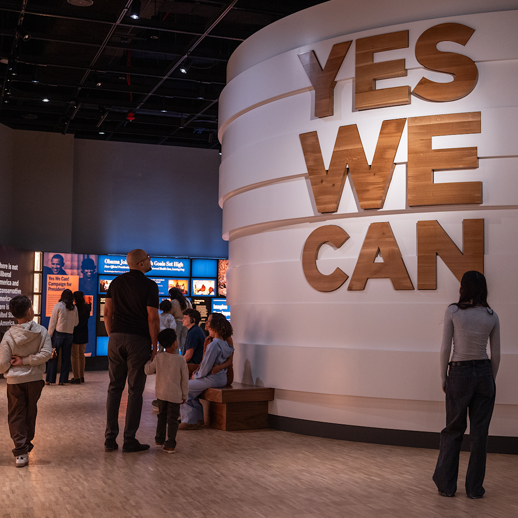 People walk by the “Yes We Can” exhibit inside the Museum Building. The exhibit is a large circular structure with words that say “Yes We Can” in wooden letters. An exhibit screen is in the background.