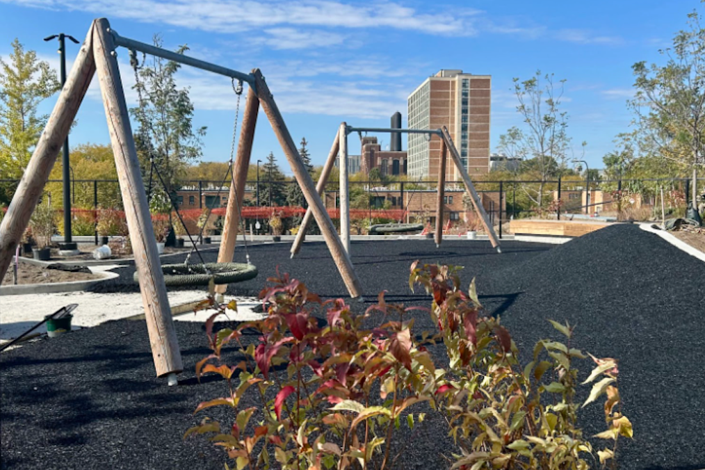 A photograph of the Obama Presidential Center Playground. The image features several swing tires sitting on top of a pebble surface. In the background of the swings are several buildings in the distance. It is daylight outside.