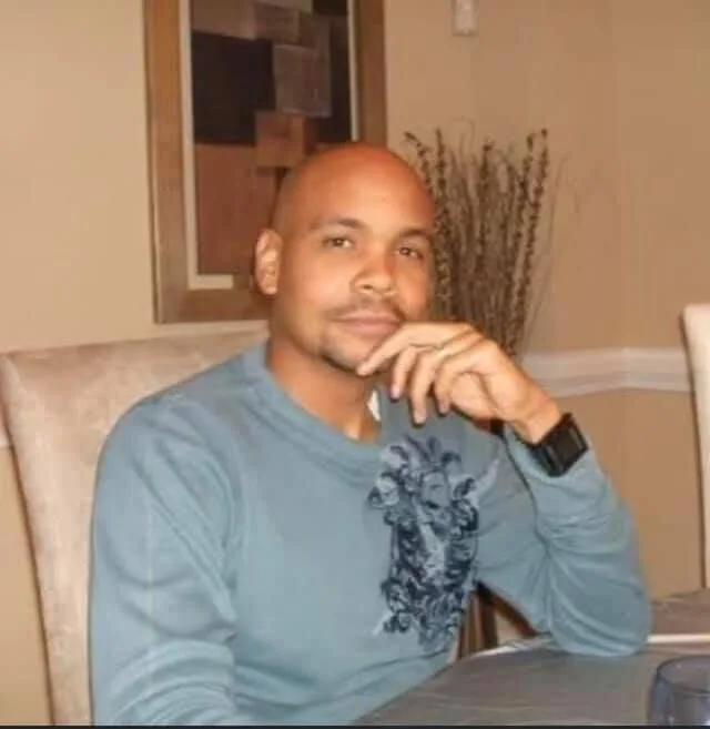 Roderic Walker, a Black man with a medium skin tone, stares into the camera as his hand rests on his cheek. He has no hair and is wearing a gray sleeved shirt.