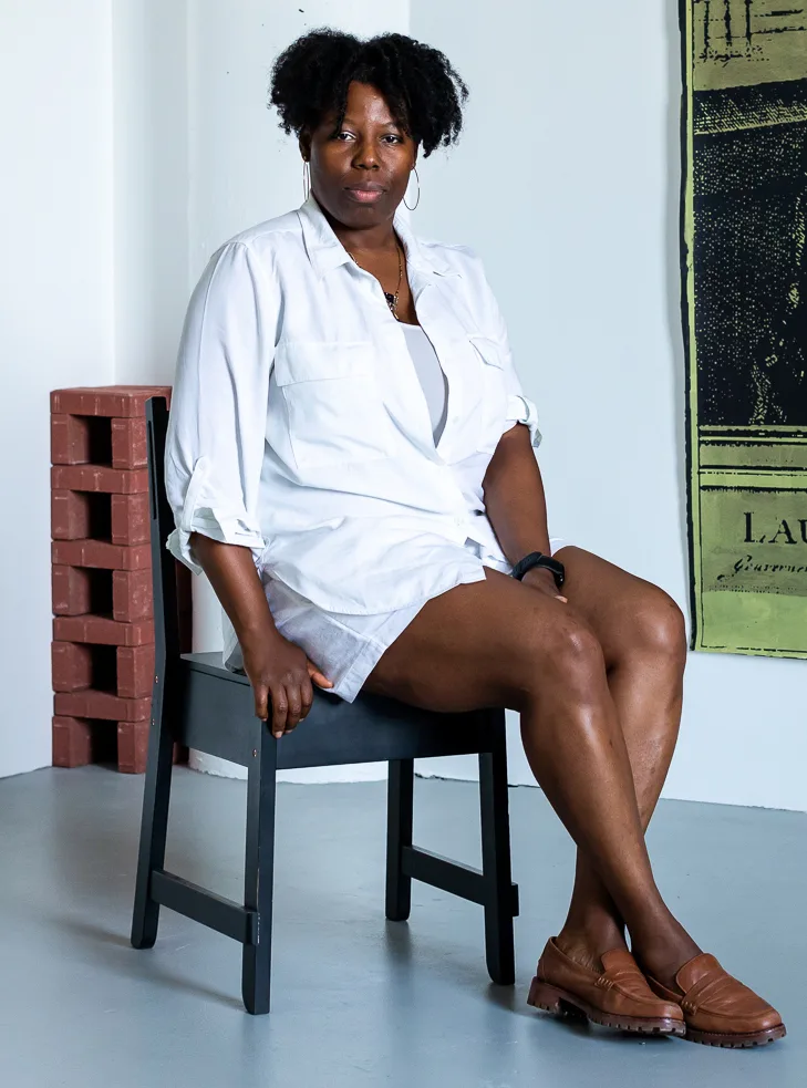 Tyanna is sitting with her legs crossed on a dark colored chair staring into the camera. Behind her is a white wall with a green tapestry hanging from one side. Next to the wall is a small stack of bricks. Tyanna has a deep skin complexion and medium length black hair. She is wearing hoop earrings, a white long sleeved shirt and a white skirt. On her feet are brown loafers.