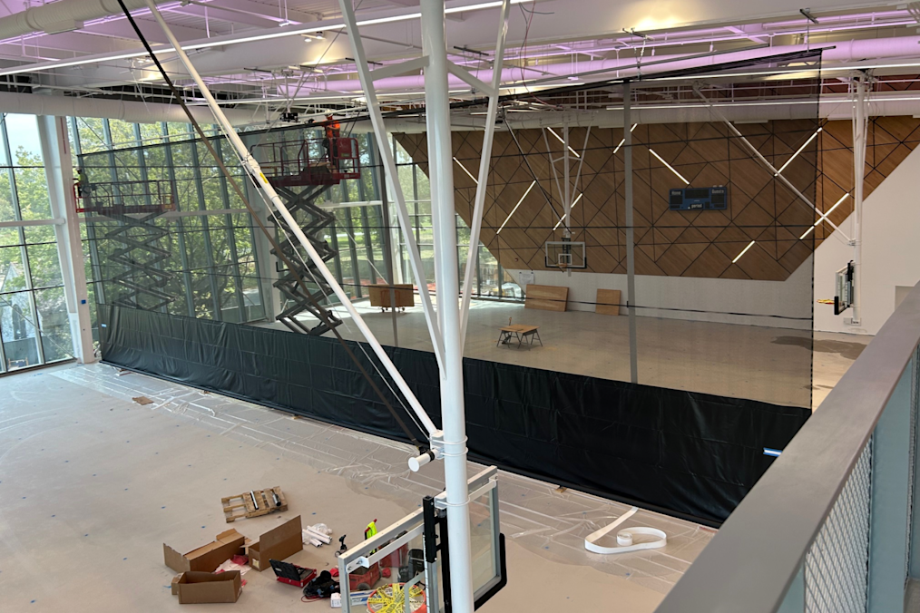A photograph of Main Court, a gymnasium located in Home Court on the Obama Presidential Center campus. The gymnasium features a wooden backboard in the shape of a basketball net. In the middle of the court is a large black screen separating two sides of the gym. On the floor of the gym is a red crane.