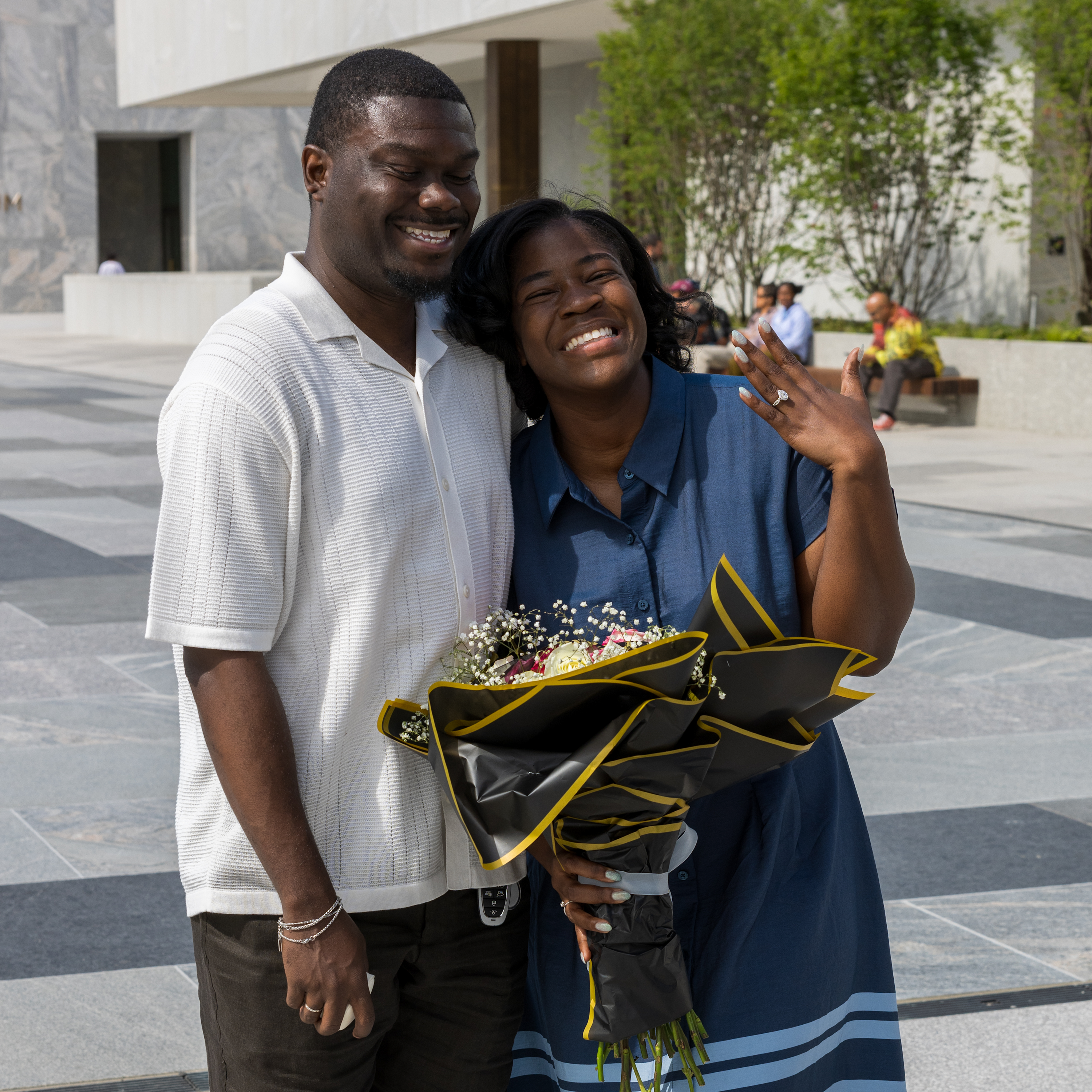 Vannessa Beene, a Black woman with a dark skin tone smiles as she holds up her left hand. She is posed next to her fiancé, Purvis Funches, a Black man with a dark skin tone. They are smiling as they stand on the John Lewis Plaza at the Obama Presidential Center.