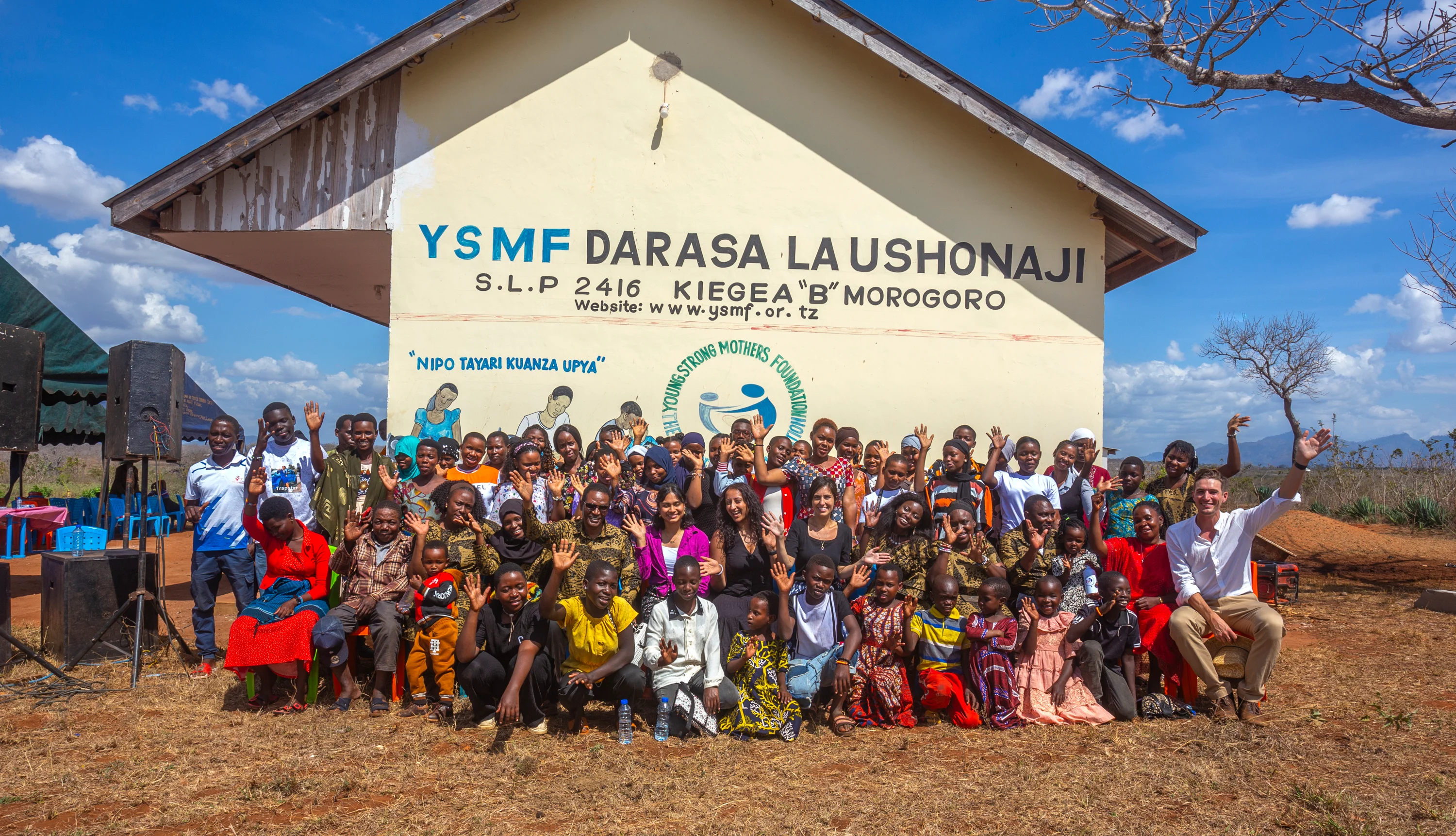 A group of girls with a range of ages and light to dark skin tones gather in front of a building on a sunny day. The side of the building reads, “YSMF Darasa LA USHONAJI,” which translates to YSMF sewing class.