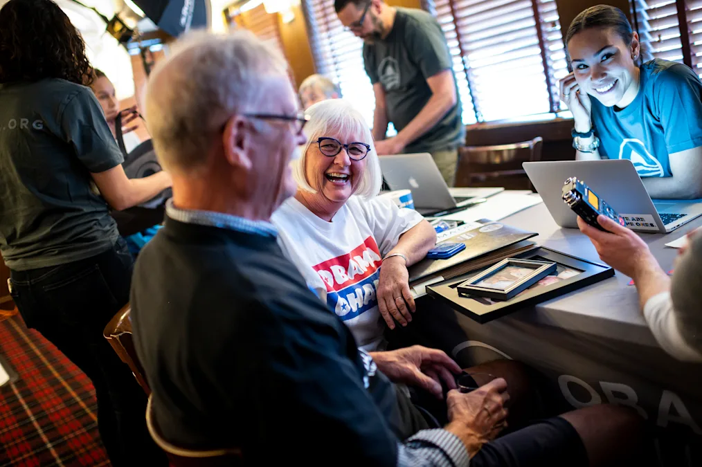 A white woman with short white hair smiles largely while resting her arm on a table. She is wearing black glasses and wearing an Obama shirt. To her right, a white man smiles as he speaks into a tape recorder. His side profile is facing the camera.