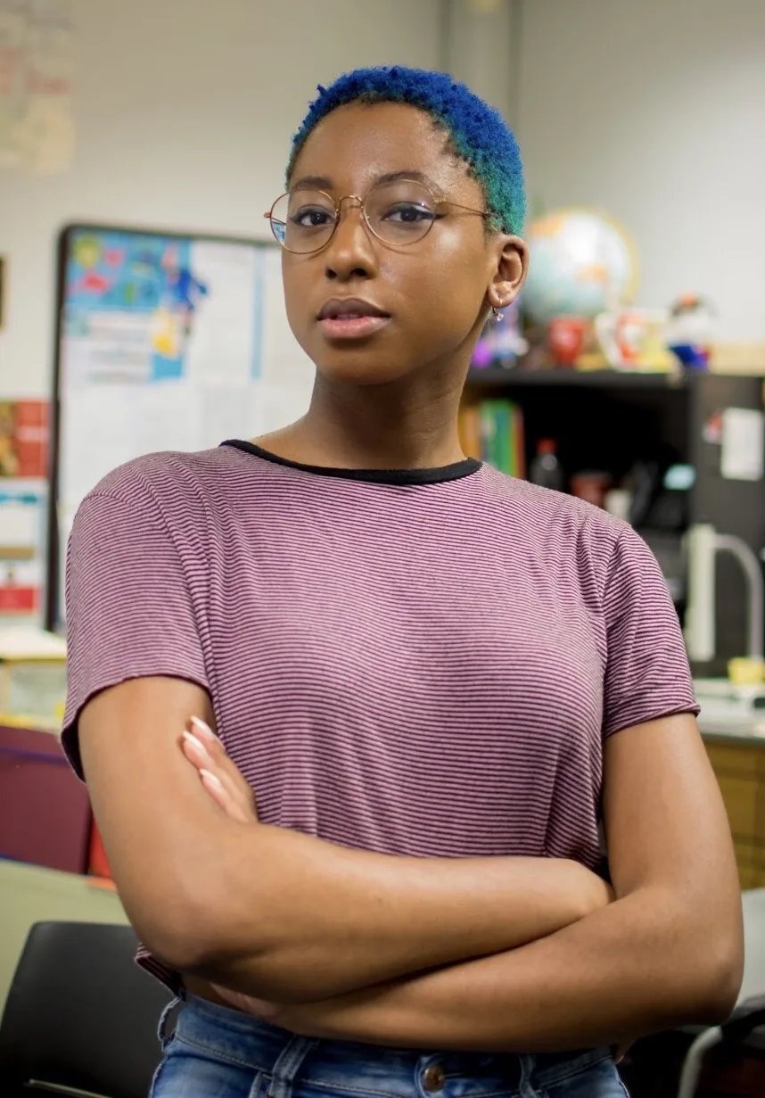 A medium skin toned Black woman with blue, close cut hair is standing with her arms crossed look at the camera with a straight expression. She is wearing gold framed round glasses, a red and black striped t-shirt and an earring in her right ear. The background is a classroom. 