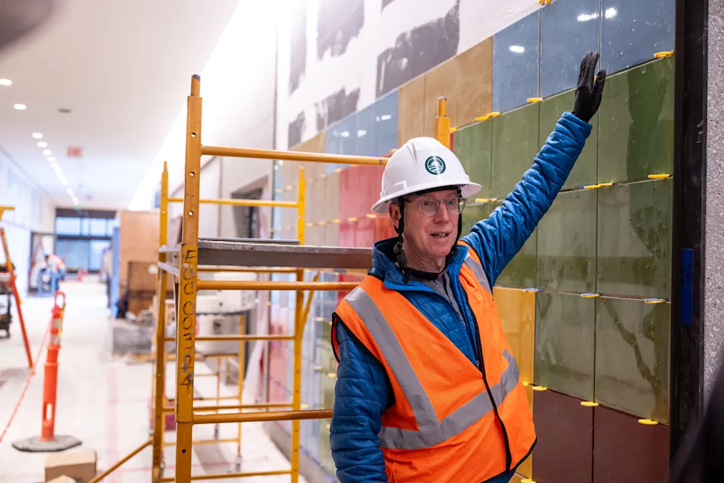 Artist Spencer Finch standing next to his artwork, To Disappear Enhances – (Memory Landscapes).” Spencer has a pale complexion. He is wearing glasses, a white hard hat, black work gloves, an orange construction vest, and a blue puffer jacket. Behind him is a yellow crane.