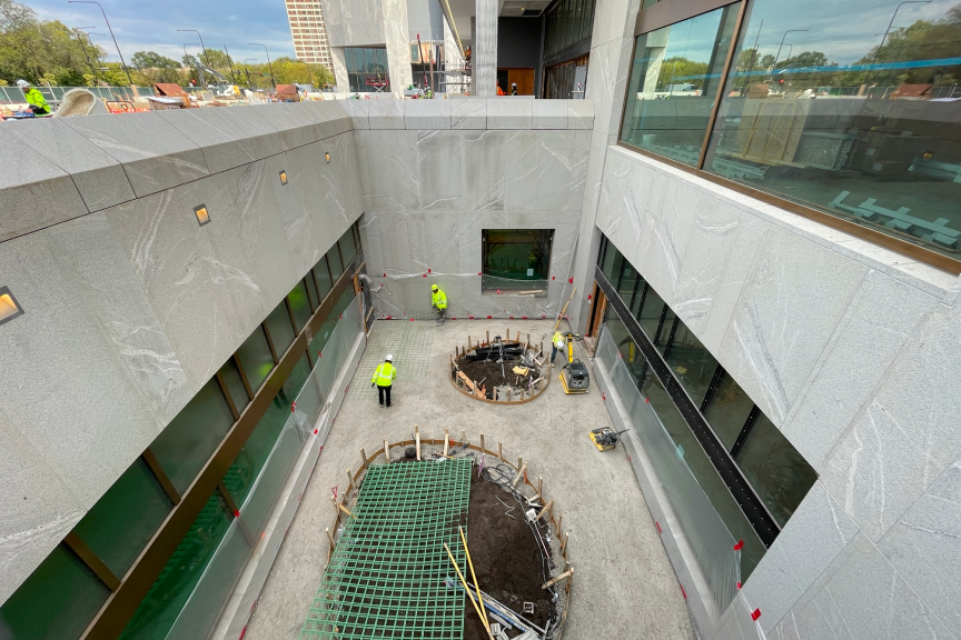 The photograph is a look at one of the outdoor Courtyards on the Obama Presidential Center campus. The picture is taken from above. Below is a circle of dirt covered by green netting. There are several construction workers standing in the courtyard area. Surrounding the courtyard are several windows and concrete.