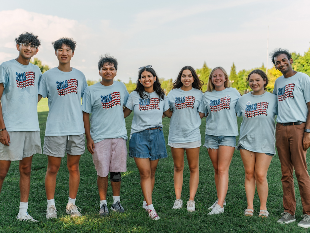 Eight smiling young people stand shoulder-to-shoulder on green grass outside. They all wear matching light blue t-shirts with "2018 NEW VOTERS" printed across the front in red, white, and blue. The group has diverse skin tones, ranging from light to medium-dark. Behind them are green trees and a soft sky.