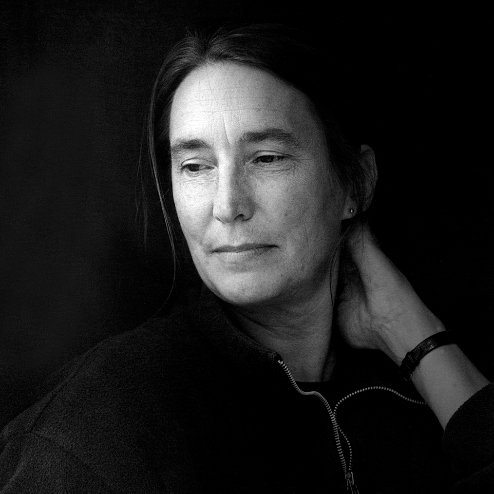 The image is a photographed portrait of Jenny Holzer. The image is in black and white. Jenny has a pale complexion and long dark hair. She is looking off into the distance and touching her hair with her left hand. She is wearing a black jacket with a silver zipper that is zipped up to her neck.