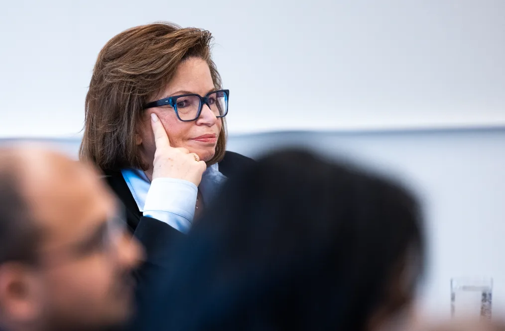 Valerie Jarrett sits listening with her hand up to her face. She wears blue and black glasses, a light blue blouse and a black jacket.