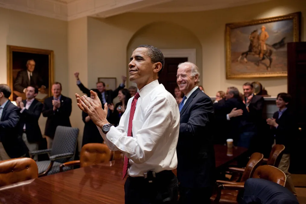 President Obama and then Vice President Joe Biden clap as they stand at a conference table. In the background, people with a range of light skin tones clap and cheer. All are dressed professionally. 