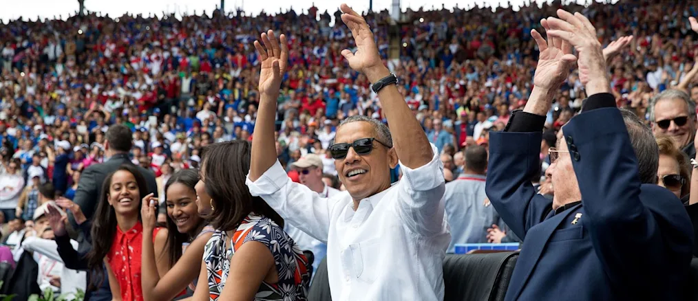President Obama is wearing sunglasses and a white collared shirt raising his arm toward the camera. The background includes the first family and a large crowd behind them.