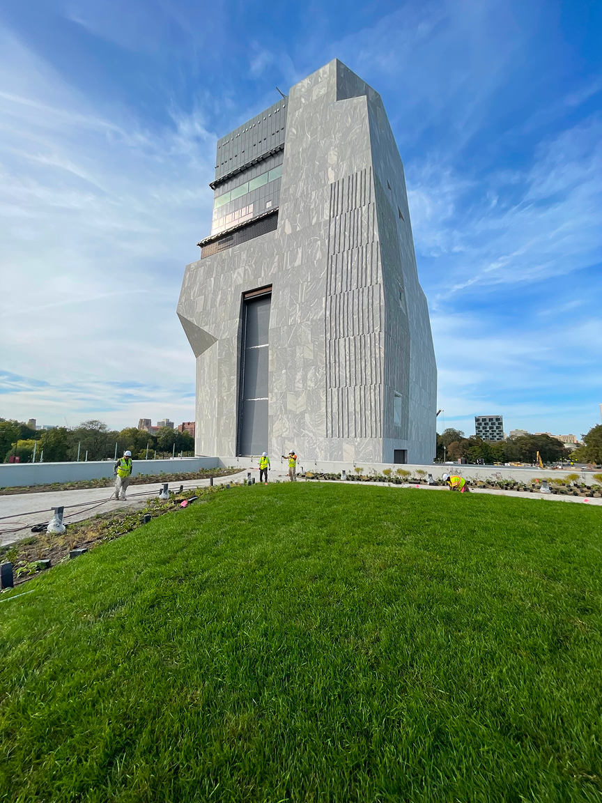 A photograph of the exterior of the Obama Presidential Center Museum. The Museum is a tall gray building. In front of the building is a plot of green grass. In the background is blue sky.