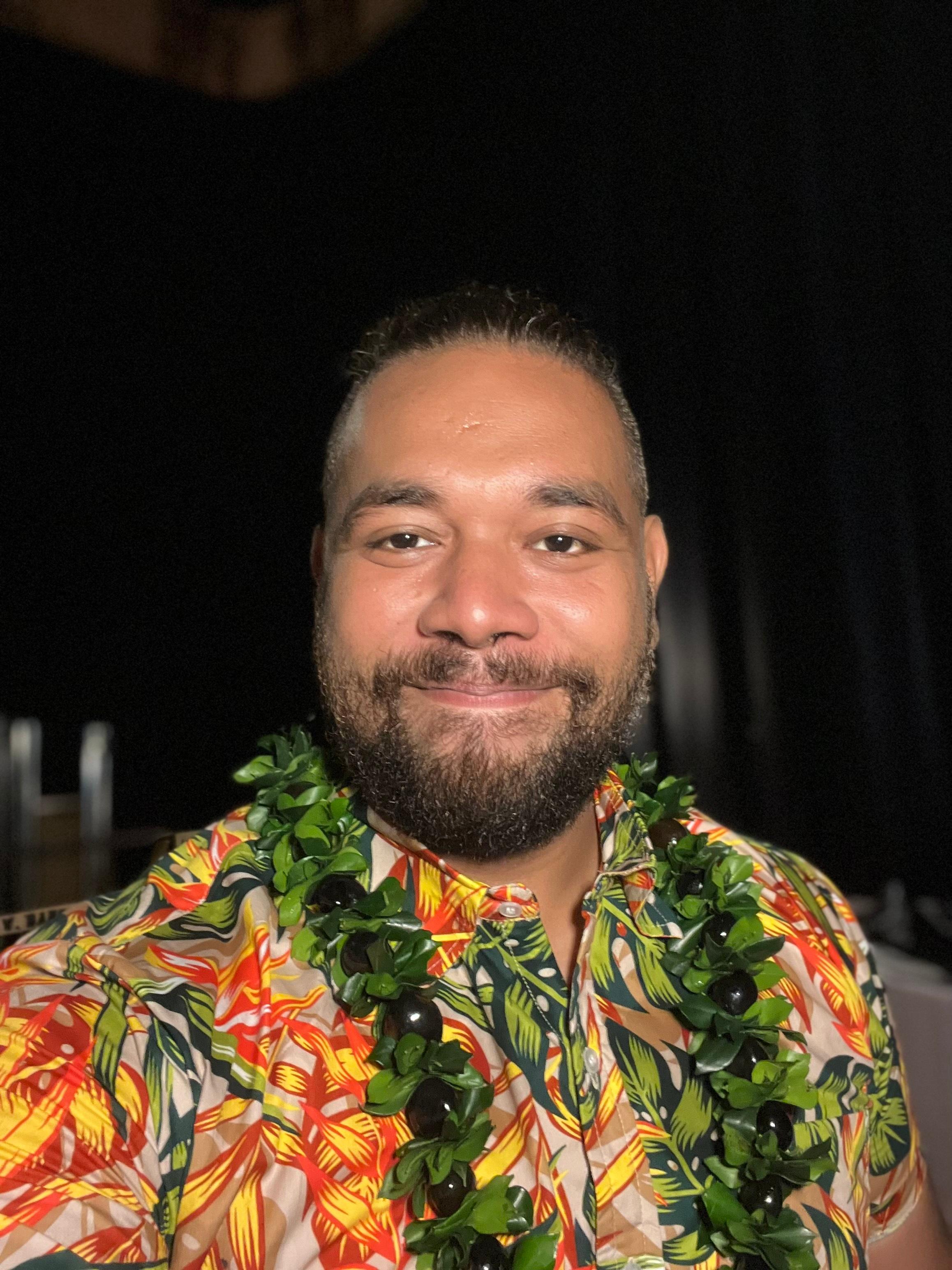 John Taukave, a man with a medium skin tone, holds a closed lip smile. He has dark hair and a beard. He is wearing an orange and yellow patterned shirt with a green lei.