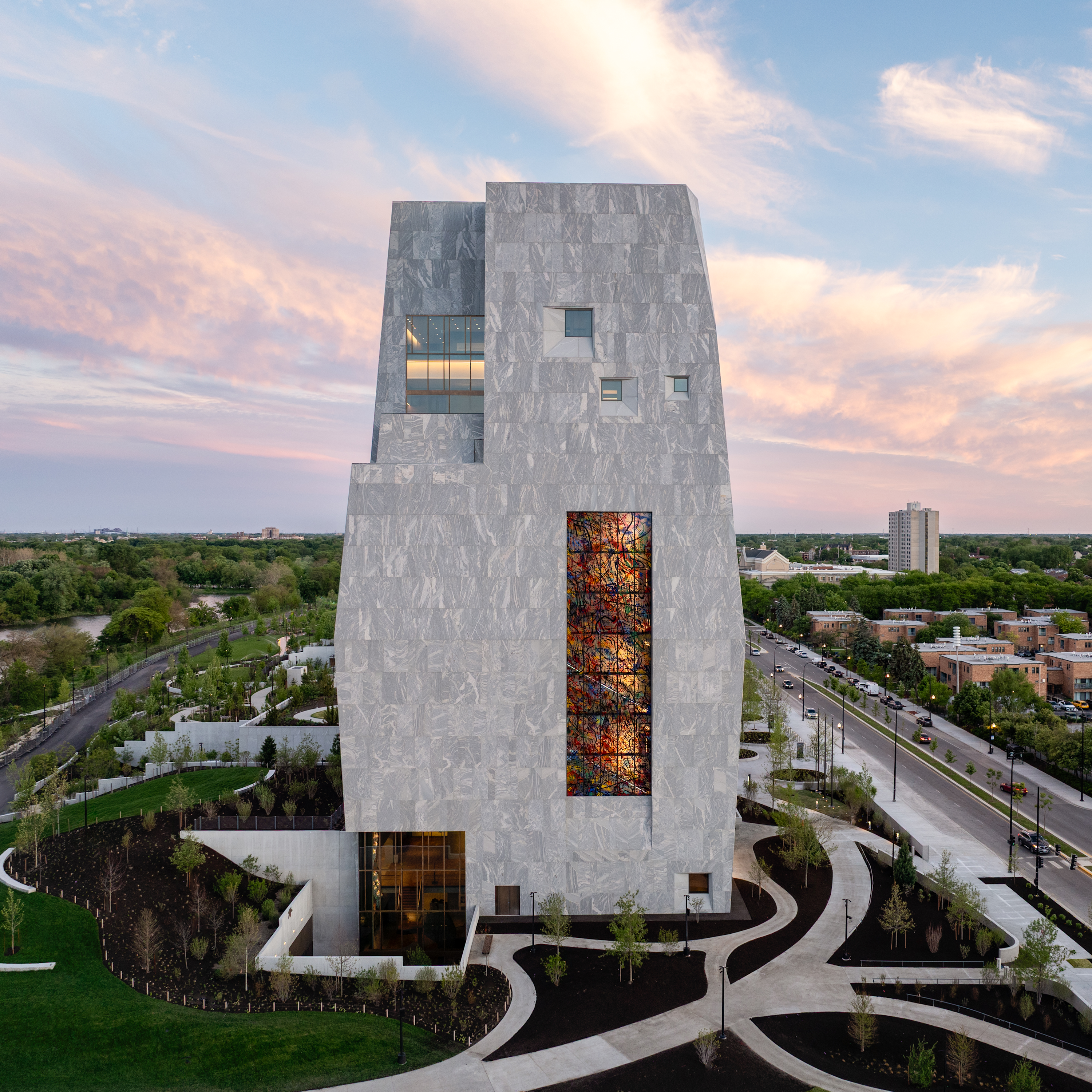 A high-angle photo shows the Obama Presidential Center Museum which is a tall light gray stone building on a green lawn during sunset. The building has an uneven shape with small windows. A tall rectangle filled with colorful stained glass sits near the center of the tower. At the bottom left, large glass windows light up an entrance. Smooth concrete paths curve around black dirt gardens and young green trees in front of the building. On the right, a multi-lane street with cars runs alongside a row of buildings and trees. In the background, green trees stretch toward the horizon under a soft pink and blue sky with light clouds.