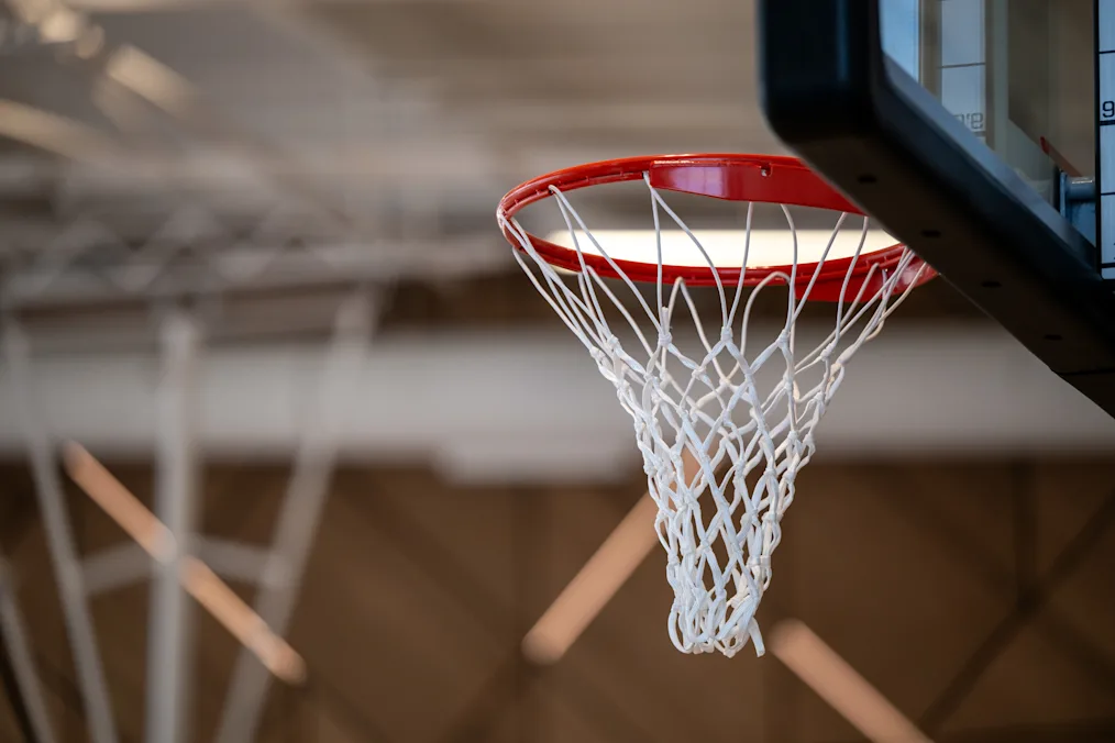 The image is a close-up shot of a basketball net hanging on Main Court located in Home Court at the Obama Presidential Center. The basketball net is hanging from a black backboard. The net is white and the rim is red.