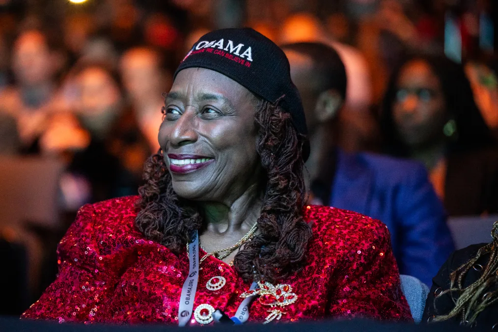 A Black woman with a deep skin tone smiles away from the camera. She is wearing a red sequin top and a hat that reads, “Obama, change we can believe in.” 