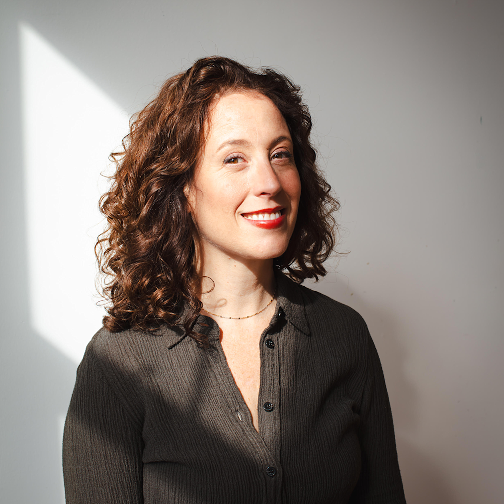 A photographed portrait of Aliza Nisenbaum. Aliza is sitting in a black chair that is partially obscured in a room with a white wall. Alize has an olive complexion and long dark curly hair. She is smiling at the camera. She is wearing a small dark necklace and a dark colored button-down shirt.