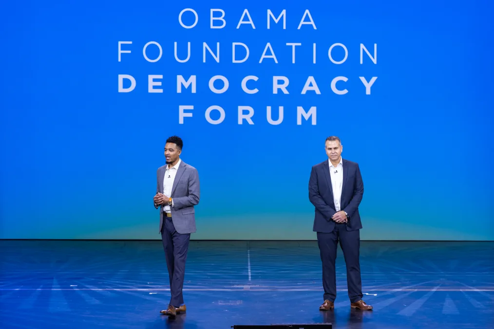  DeMario Phipps-Smith, a Black man with a medium skin tone stands next to Peter Adams, a man with a light skin tone on a stage. The screen behind them reads, “Obama Foundation Democracy Forum.”