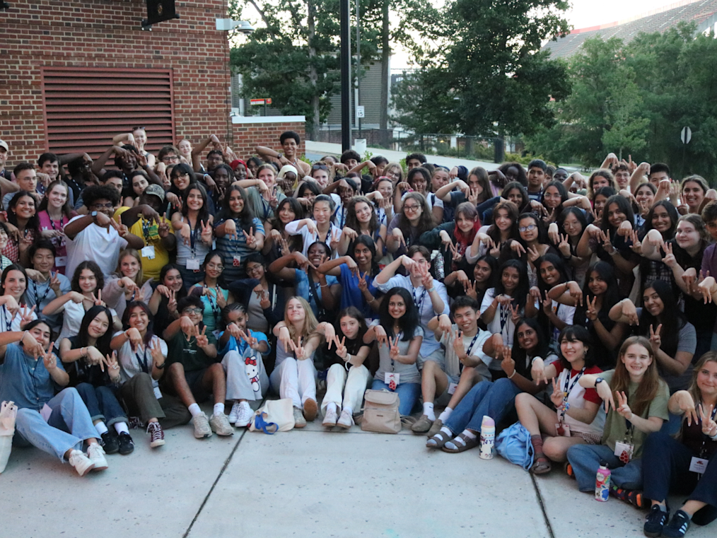 A large, diverse crowd of dozens of teens and young adults poses outside a red brick building. They sit on the ground and stand in rows, smiling broadly at the camera. Many hold up both hands to form an "M" shape or peace signs. The individuals have light, medium, and dark skin tones