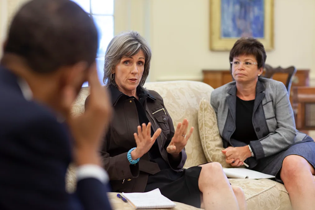 The camera focuses on an older woman in business attire with a light skin tone and gray short hair as she sits on a couch in the middle of a conversation. Beside her is a woman with a medium skin tone short brown hair and glasses in business attire