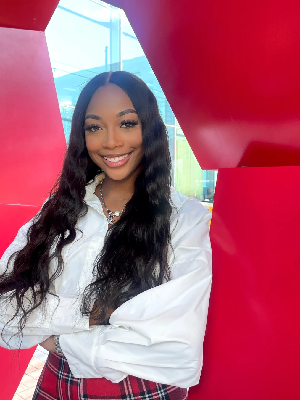 Raven Smith, a Black woman with a medium skin tone, smiles at the camera. She is wearing a long sleeved white shirt and red plaid pants. Her arms are crossed and she has long black hair.