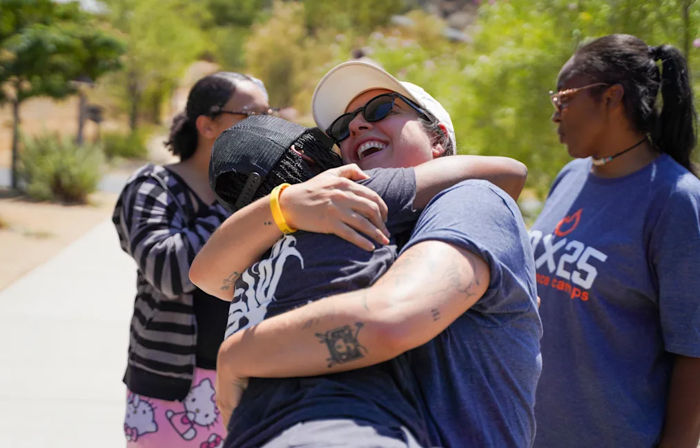 Jesse Moss, a woman with a light skin tone, wraps her arms around someone. They are surrounded by women with a range of light to dark skin tones. Jesse is wearing a white hat and black sunglasses.