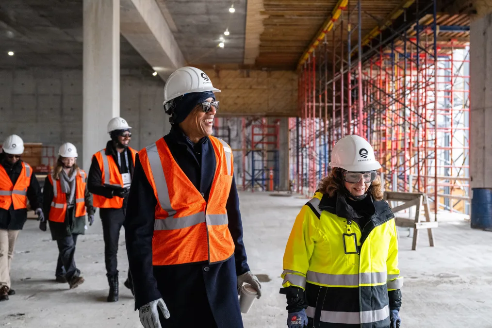 President Obama is smiling wearing sunglasses, a white hardhat with the Obama Foundation rising sun logo, gloves, and a bright orange safety vest over a long dress coat. He is holding a cup of coffee and standing next to a white woman wearing a neon yellow construction coat, hard hat, and goggles. They are walking in front of three other people with a range of light to deep skin tones wearing orange safety vests and white hard hats. They are all walking in an unfinished building with scaffolding in the background.