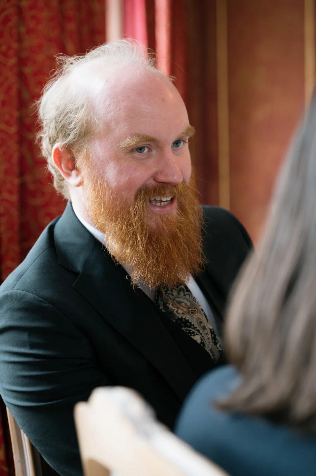Bjorn Ihler, a man with a light skin tone and thin red hair, smiles as he speaks with someone whose back is to the camera. He has a long red beard and is wearing a black suit. 