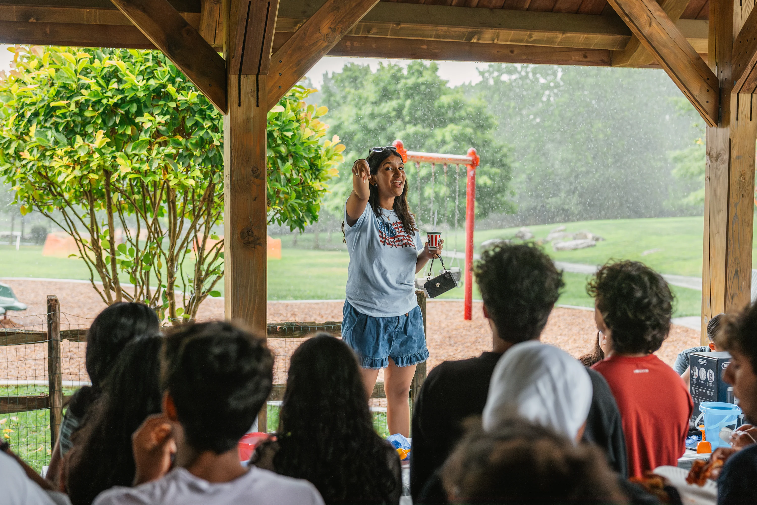 A young adult with medium skin tone stands in an open-sided wooden pavilion and points forward while speaking. They wear a light blue t-shirt and denim shorts. In front of them, a group of listeners sits with their backs to the camera, looking toward the speaker under falling rain.